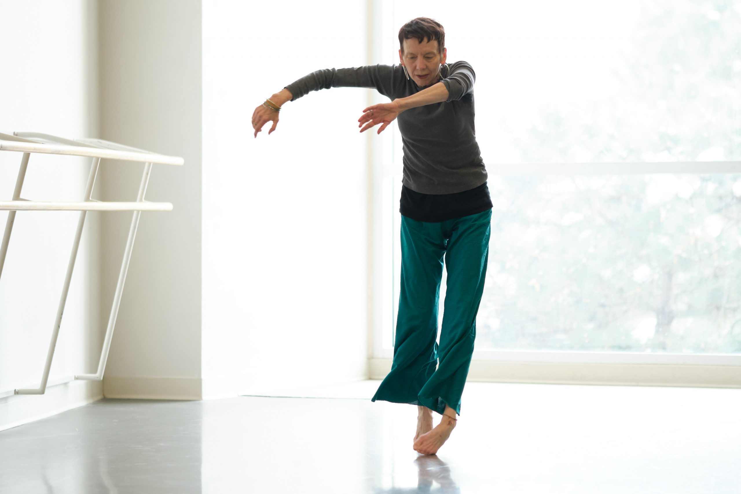 A light-skinned dancer in her 60s tosses her arms in a curve to the left while she rises on her toes