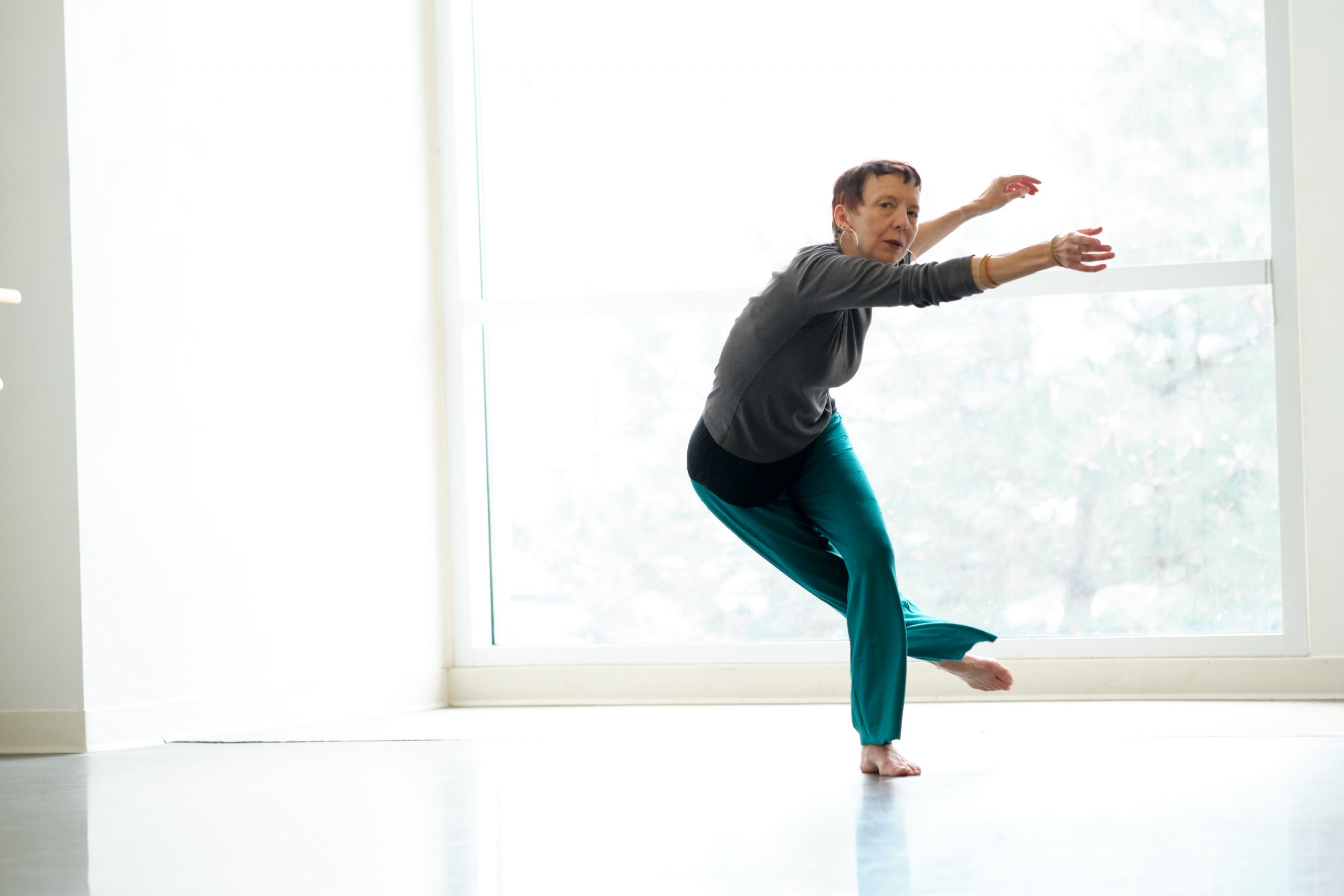 Erin, a light skinned dancer in her 60s, tosses both arms and one leg to the left as she leans to the right