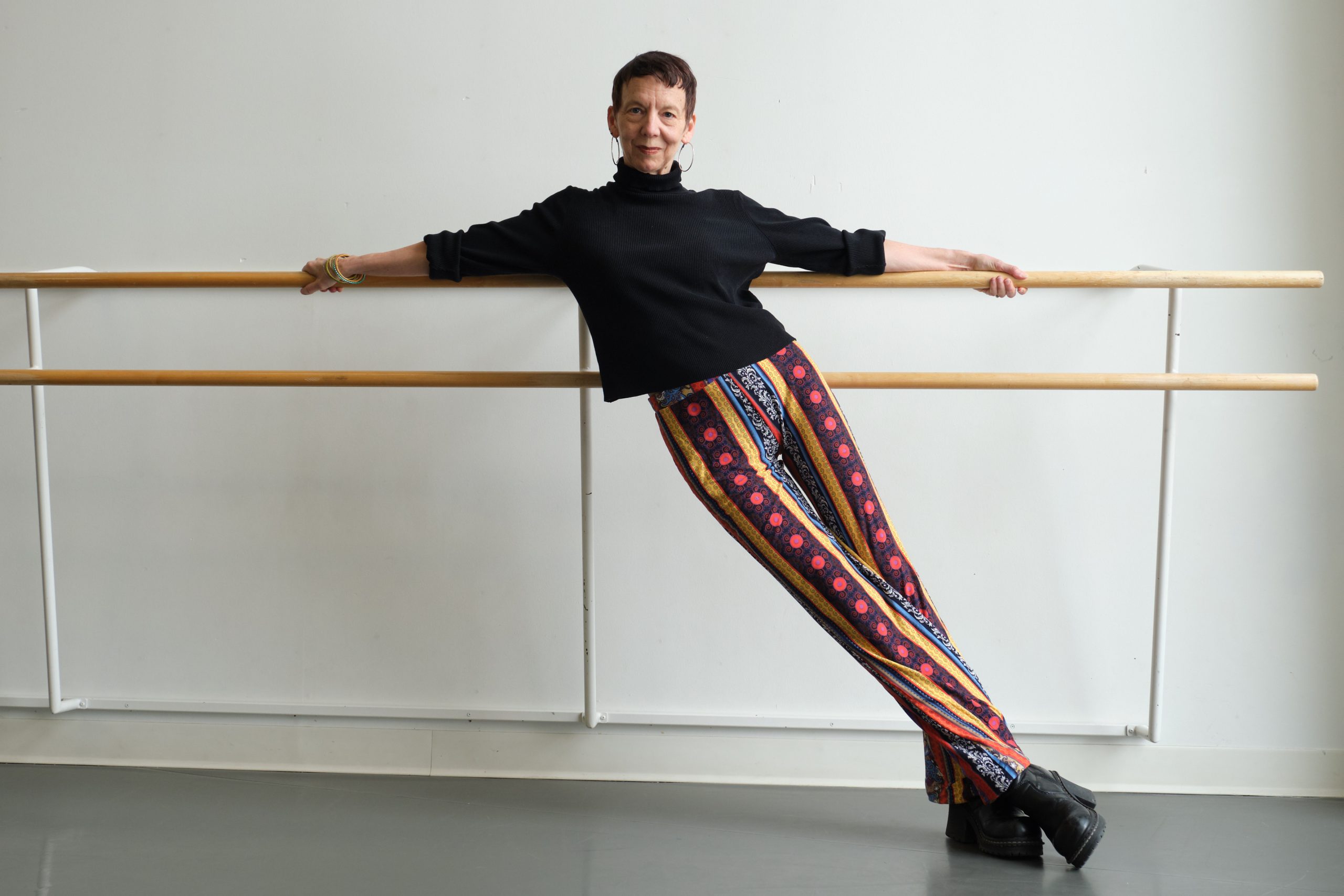 A light-skinned woman in her 60s, with close cropped hair, leans against wall-mounted ballet barres and tilts off to the left