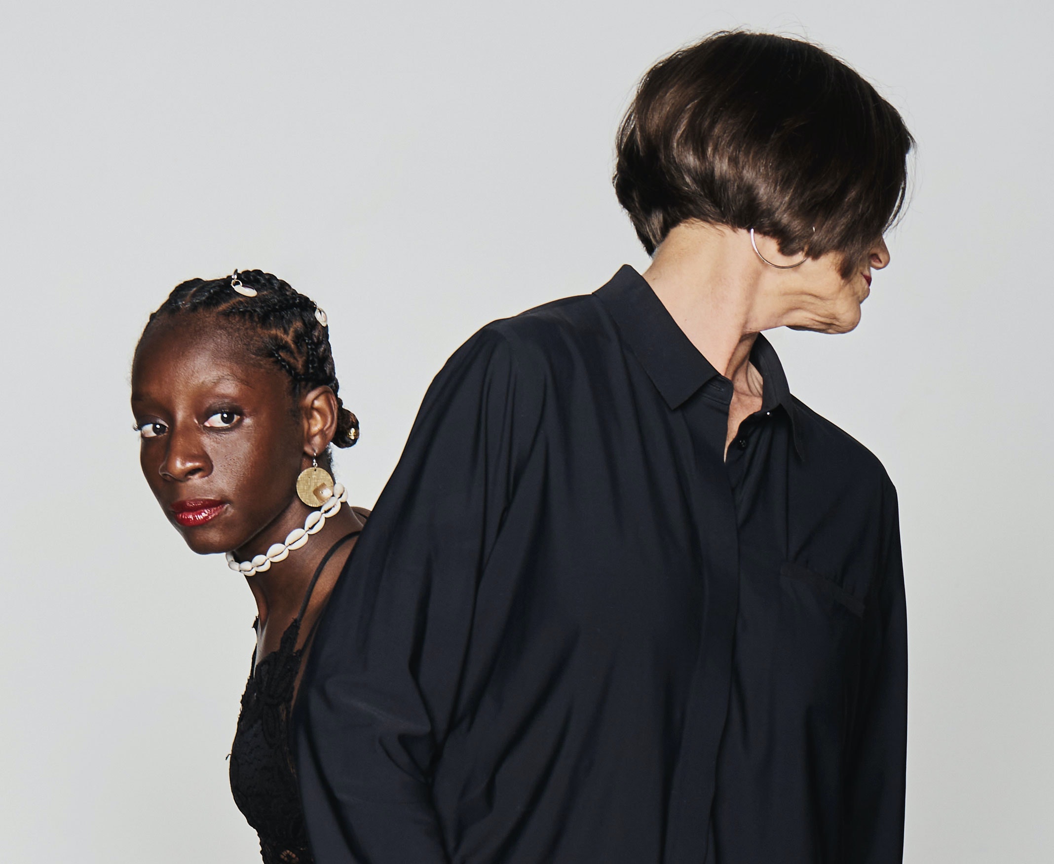 Judith Howard, a tall white woman stands near the camera, her face is turned over her shoulder. Alanna Morris, a small black woman, stands behind Judith and looks around Judith to face the camera