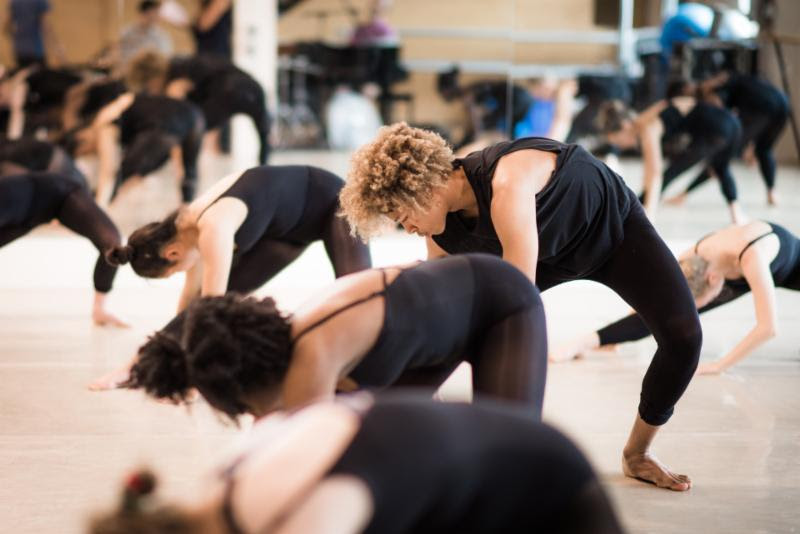 A collection of dancers, many of them black, lean over to their left sides like gathering something from the earth