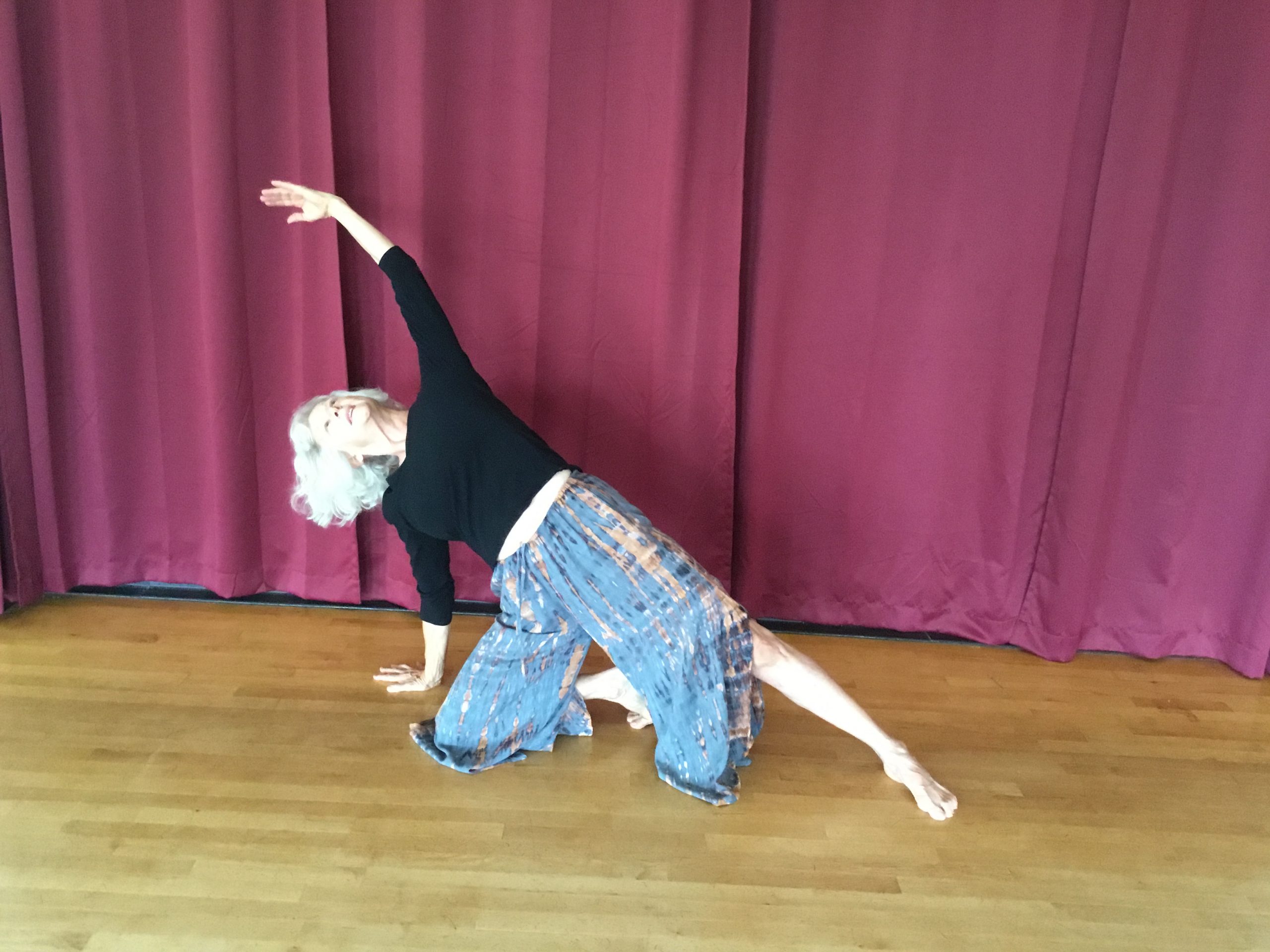 A light-skinned dancer with white hair supports herself on one hand and one knee, with the other arm and leg outstretched