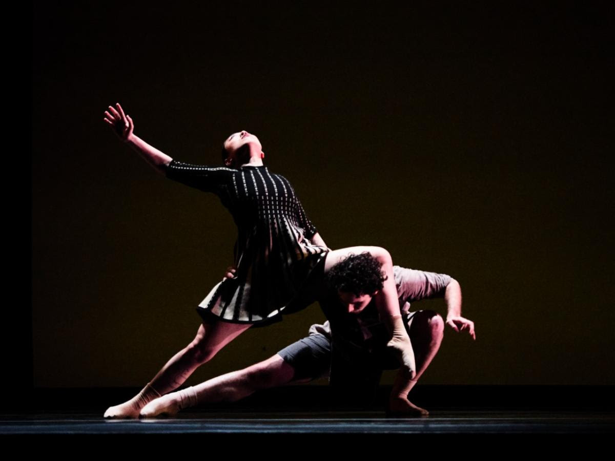 Two dancers one a dark stage perform deep lunges in dramatic lighting