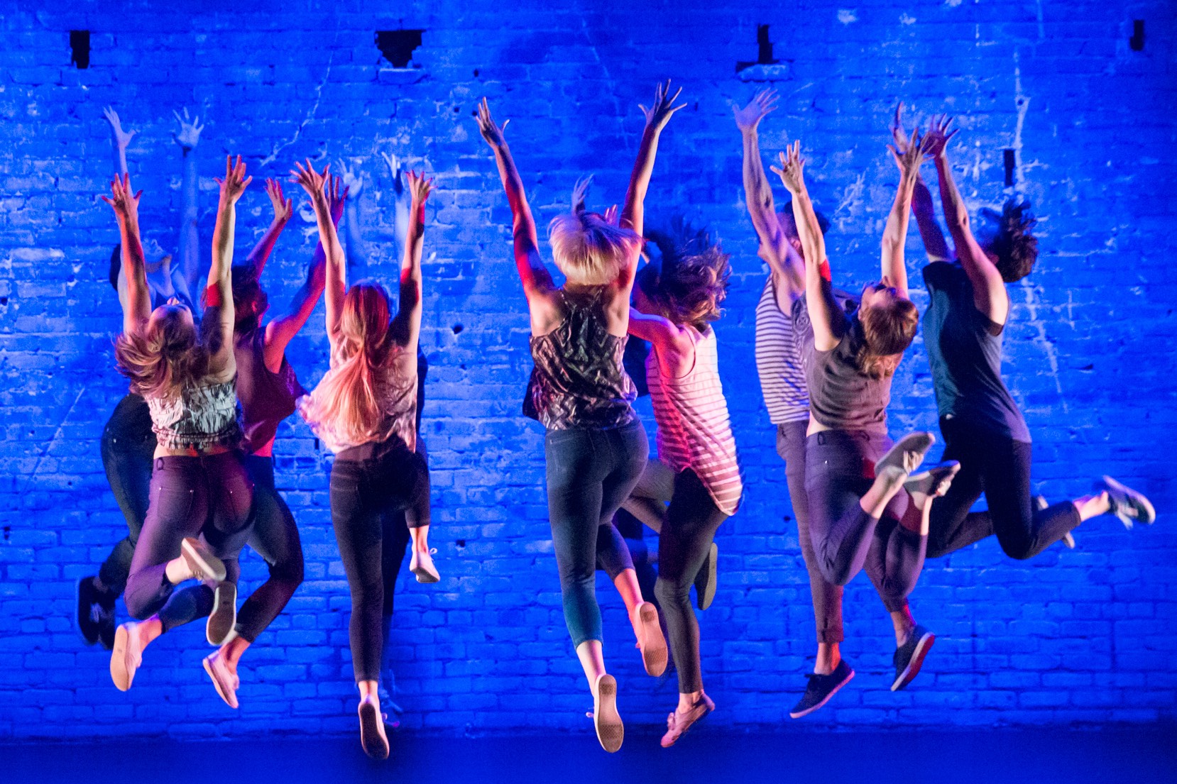 a group of dancers forming a circle are pictured mid-jump, arms overhead