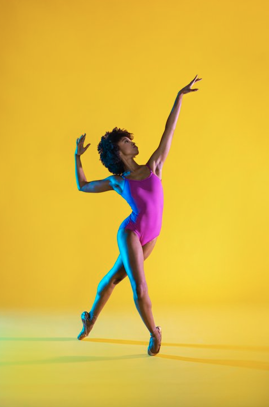 A dark-skinned dancer with a loose afro stands en pointe in plie tendu derrier as she arches back and gestures with her arms