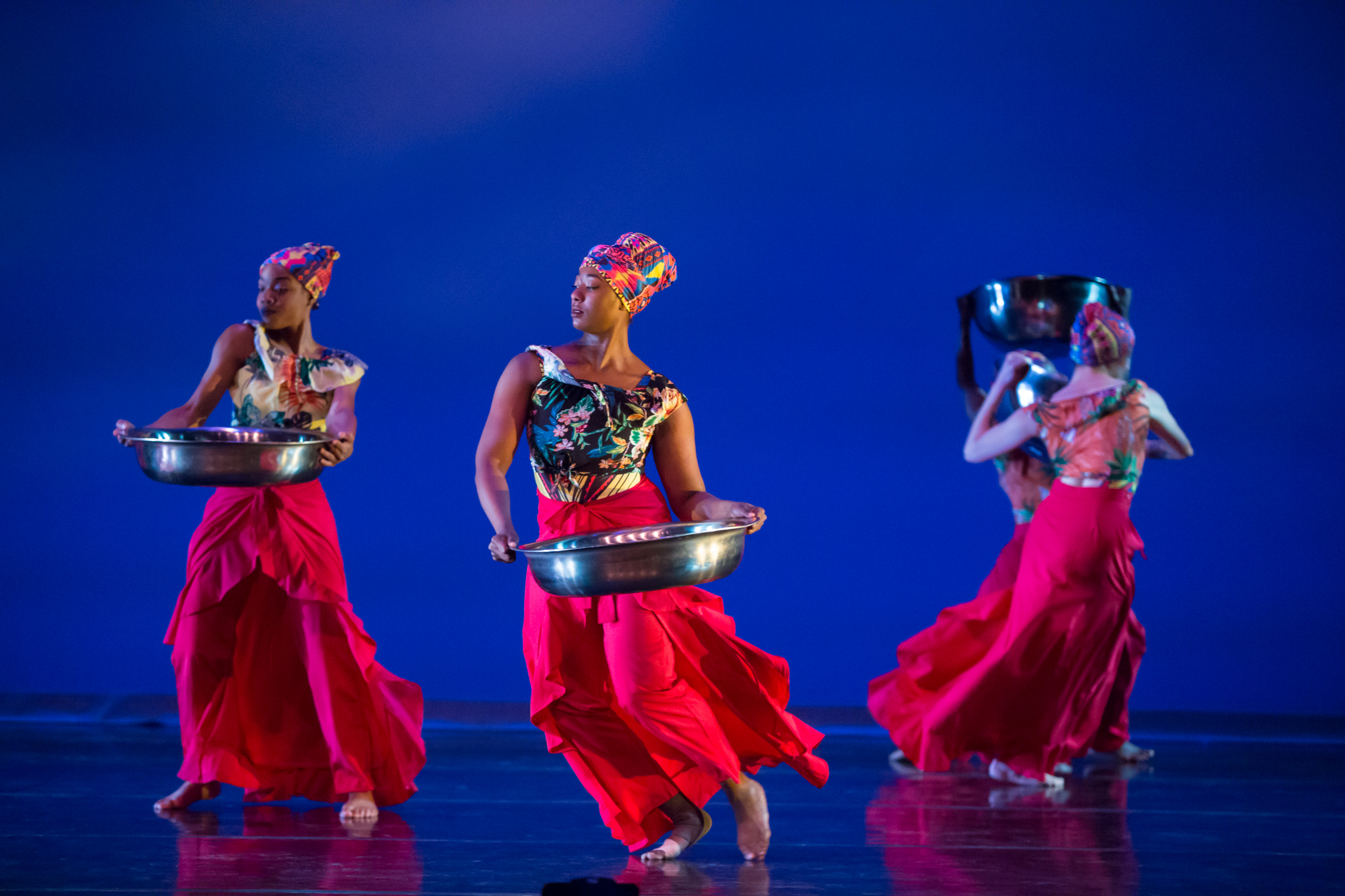 Three dark-skinned dancers in rich red skirts and head wraps move swiftly across a stage carrying a basin