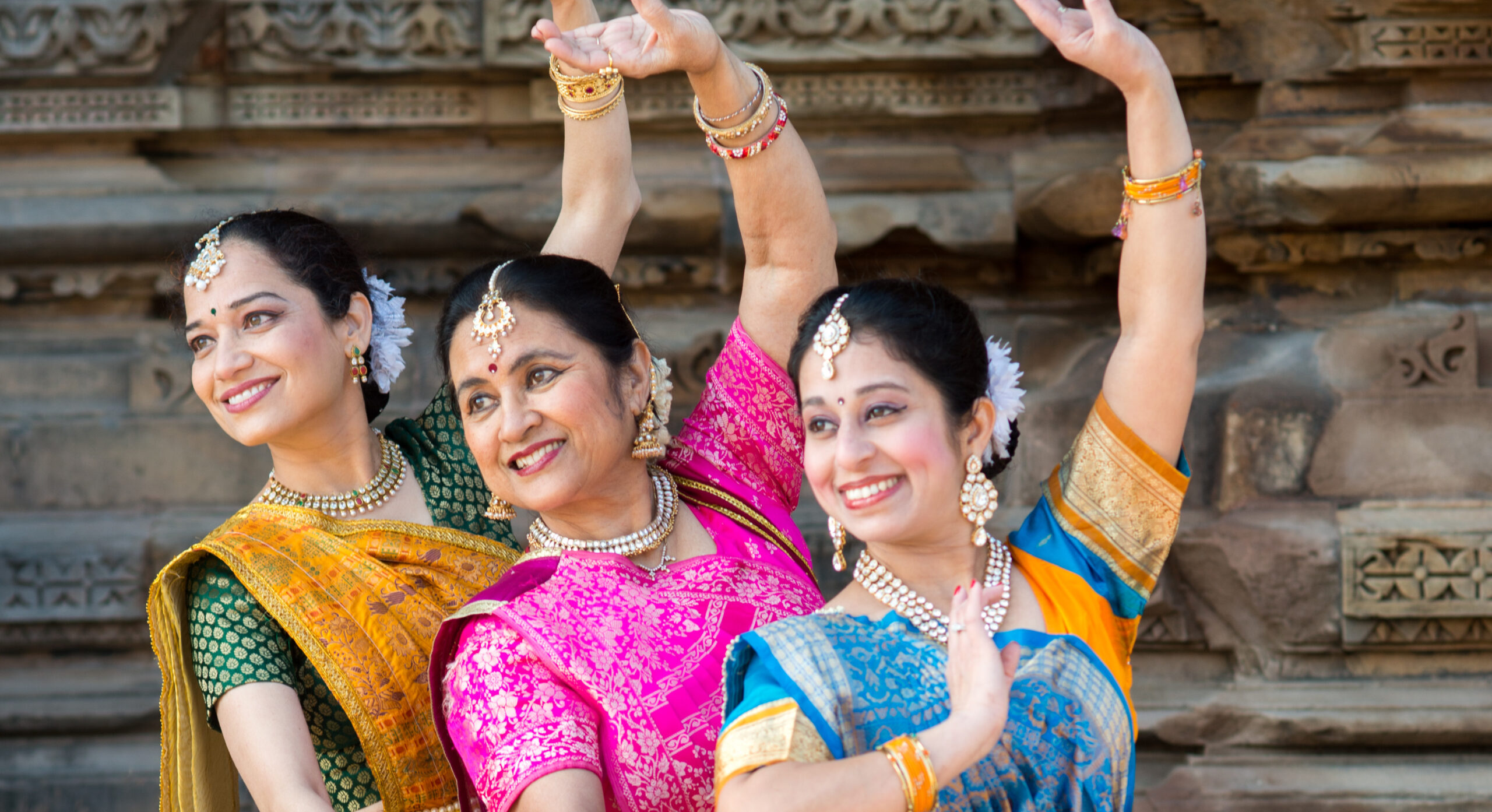Three women in traditional classical Indian dance dress smile and pose with one arm over head