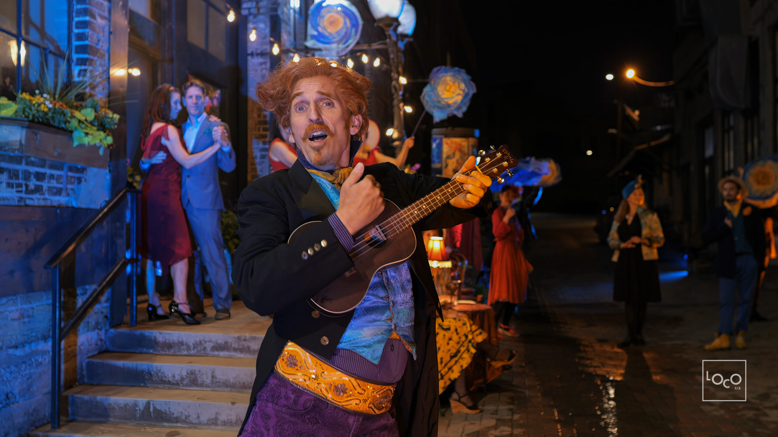 A light-skinned man with facial hair, and ornate jacket and costume, and string instrument gazes past the camera with a whimsical expression