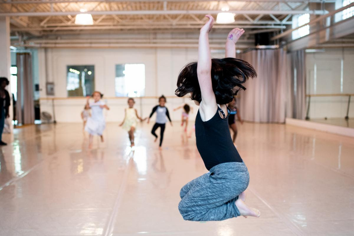 A dancer in the forefront jumps, with arms thrown over head and legs tucked in. In the background, several young dancers watch