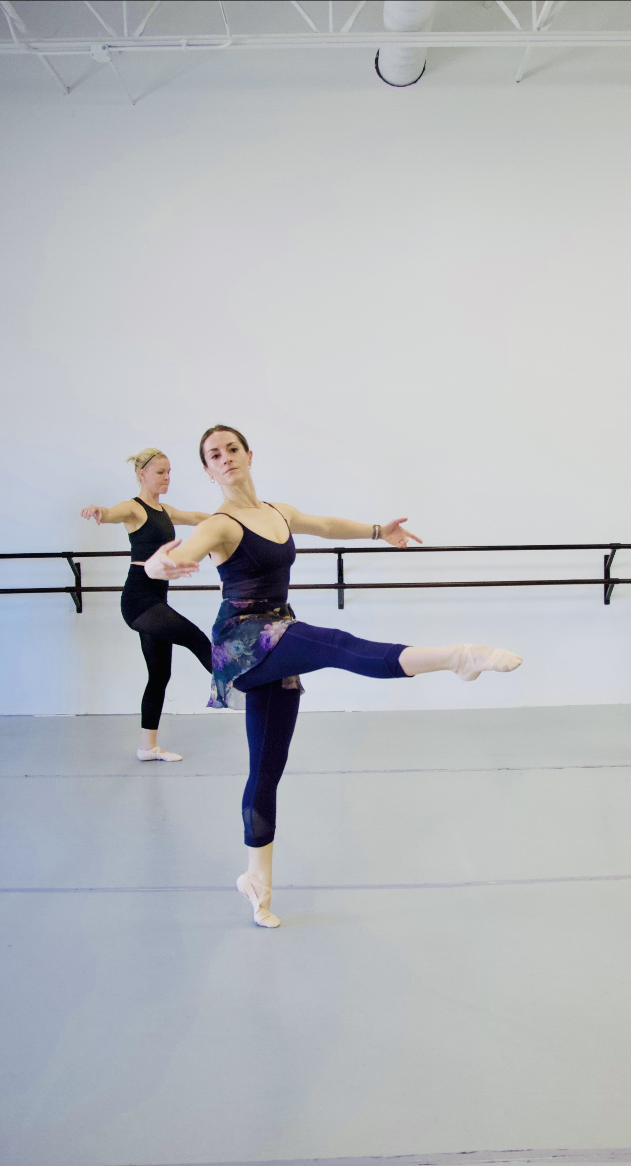 Two adult dancers in a ballet studio stand in attitude front with arms outstretched to the sides